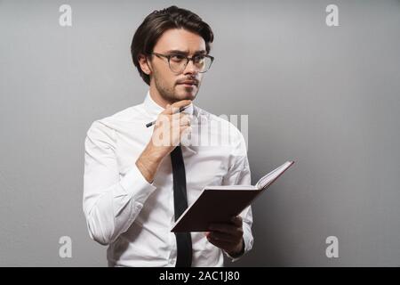 Portrait of a handsome pensive young businessman wearing suit standing isolated over gray background, holding a journal Stock Photo