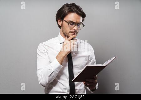 Portrait of a handsome pensive young businessman wearing suit standing isolated over gray background, holding a journal Stock Photo