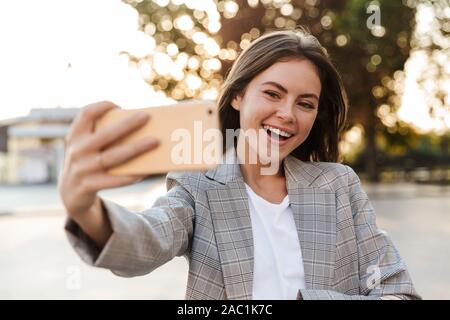Photo of a smiling positive young business lady walking by street take selfie by mobile phone. Stock Photo