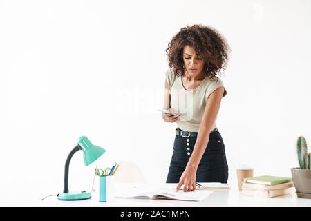 Beautiful concentrated young african businesswoman wearing casual clothing standing at the desk isolated over white background, using mobile phone Stock Photo