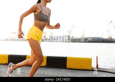 Image of energetic asian woman in sportive clothes running while doing workout against sea port Stock Photo