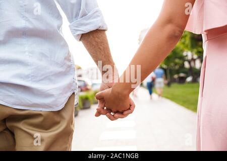 Cropped photo of a young loving couple walking by street outdoors holding hands of each other. Stock Photo