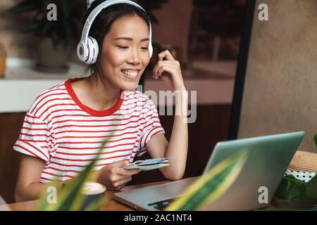 Image of a young happy cheery beautiful asian woman sit in cafe using laptop computer listening music with headphones while holding passport with tick Stock Photo