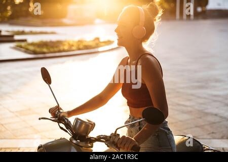 Portrait of joyful sunlit woman listening to music with wireless headphones while riding on scooter on city street Stock Photo