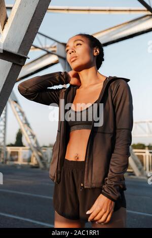 Photo of african american young woman in sportswear touching her neck while working out on old bridge Stock Photo