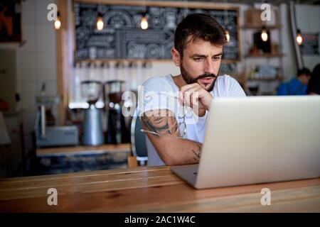 Young bearded tattooed guy in white t-shirt sitting behind the table in bar looking at his laptop Stock Photo