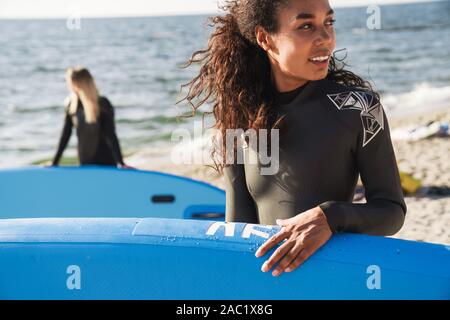 Image of joyful multinational women in wetsuits holding surfboard and smiling while working out on summer beach Stock Photo