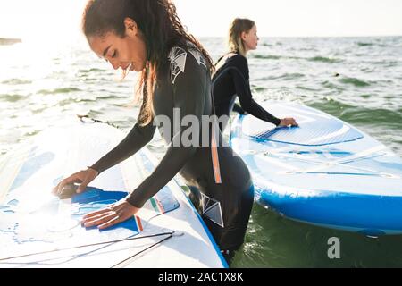 Image of happy multinational women in wetsuits using surfboard while working out on water Stock Photo