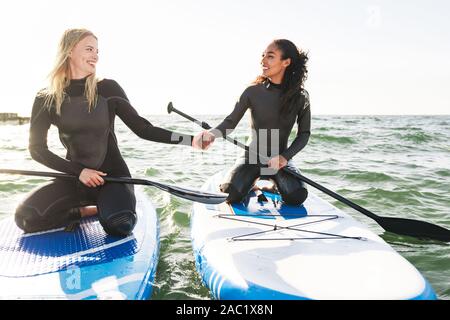 Image of joyful multinational women in wetsuits working out with surfboard and paddles on water Stock Photo