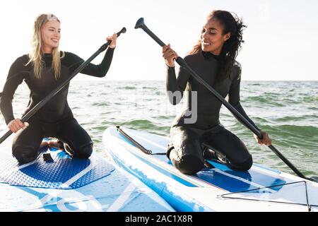Image of cheerful multinational women in wetsuits working out with surfboard and paddles on water Stock Photo