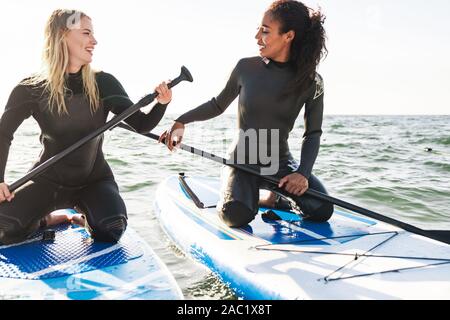Image of cheerful multinational women in wetsuits working out with surfboard and paddles on water Stock Photo