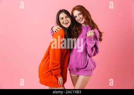 Two cheerful girls standing isolated over pink background, hugging Stock Photo