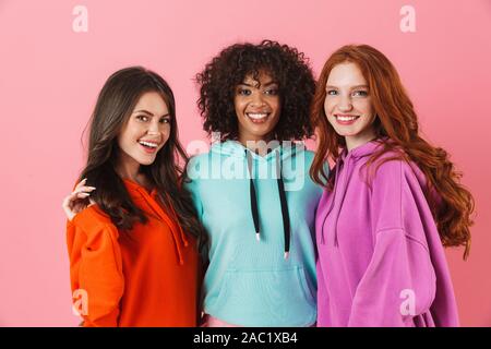 Three cheerful multiethnic girls standing isolated over pink background, hugging Stock Photo