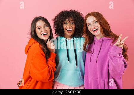 Three cheerful multiethnic girls standing isolated over pink background, hugging Stock Photo