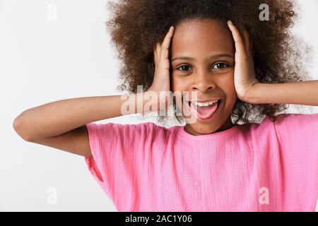 Cheerful funny little african girl wearing pink blouse standing isolated over white background, grimacing, covering ears Stock Photo