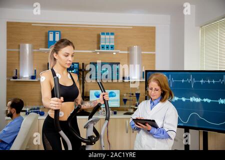 Female athlete running tests on the stepper and doctor making notes in tablet. Test with electrodes Stock Photo