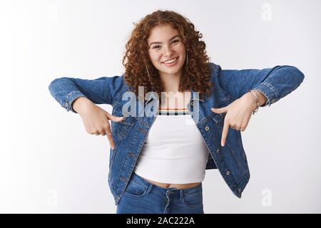 Attractive carefree young girl curly-haired redhead freckles pimples, smiling friendly raise index fingers pointing down suggesting look downwards Stock Photo