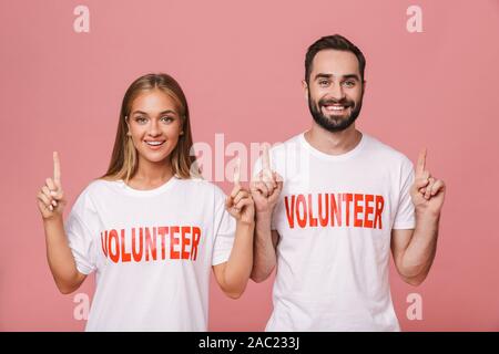 Image of beautiful man and woman volunteers wearing uniform t-shirts pointing fingers upward isolated over pink background Stock Photo