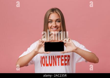 Happy young volunteer girl standing isolated over pink background, showing blank screen mobile phone Stock Photo