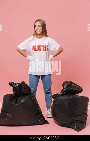 Image of cheerful woman volunteer in uniform t-shirt smiling and standing by trash bags isolated over pink background Stock Photo