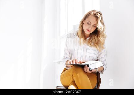 Attractive young blonde woman wearing shirt sitting on a chair indoors, reading book Stock Photo