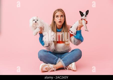 Cute lovely confused girl wearing sweater sitting with legs crossed with her pet chihuahua and lapdog isolated over pink background, choosing dogs Stock Photo