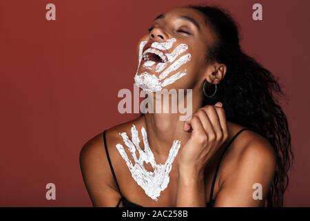Close up portrait of an attractive smiling young african woman with white hand print on her face and neck standing isolated over red background, laugh Stock Photo