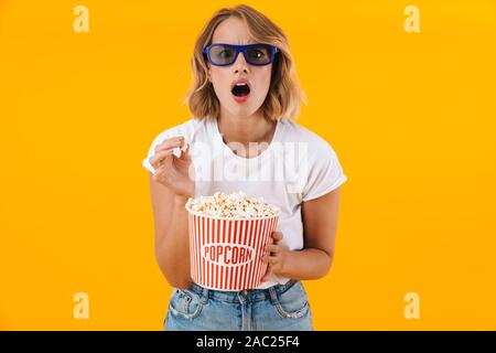 Image of shocked blond woman in 3D glasses holding popcorn bucket while watching movie isolated over yellow background Stock Photo