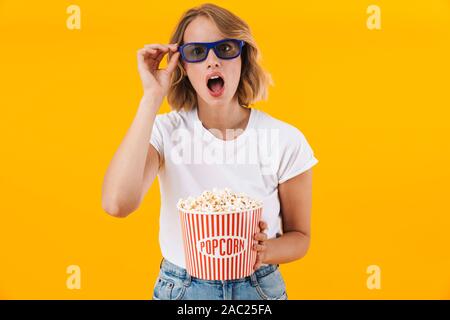 Image of excited blond woman in 3D glasses holding popcorn bucket while watching movie isolated over yellow background Stock Photo