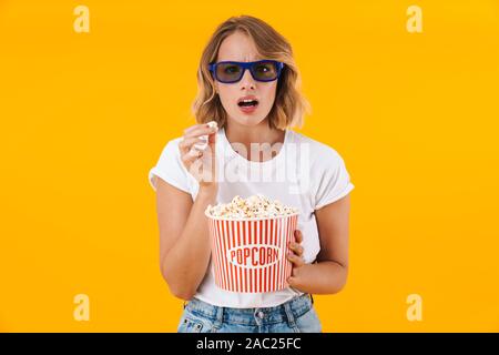 Image of confused blond woman in 3D glasses holding popcorn bucket while watching movie isolated over yellow background Stock Photo