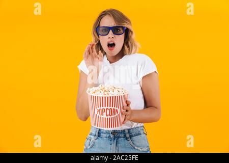 Image of scared blond woman in 3D glasses holding popcorn bucket while watching movie isolated over yellow background Stock Photo