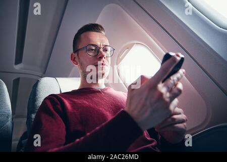 Internet connection during flight. Young man with eyeglasses using mobile phone in airplane. Stock Photo