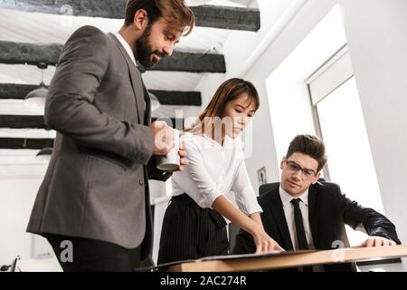 Photo of young colleagues discussing project and drinking coffee while working in open-plan office Stock Photo