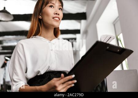 Photo of pleased asian businesswoman holding clipboard and examining paper documents in open-plan office Stock Photo