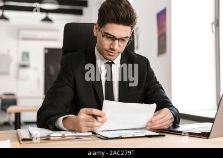 Photo of brooding businessman in eyeglasses working with documents and laptop while sitting at desk in open-plan office Stock Photo