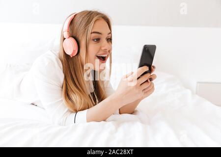 Photo of young surprised woman in pajama listening music with cellphone and headphones while lying on bed in light room Stock Photo