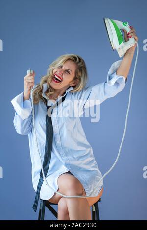 Young woman posing with an iron. Girl wearing a man's shirt and tie holding a green iron in hand. Stock Photo