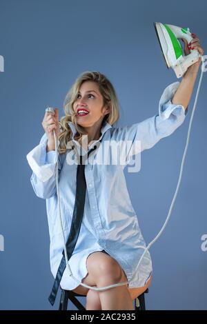 Young woman posing with an iron. Girl wearing a man's shirt and tie holding a green iron in hand. Stock Photo