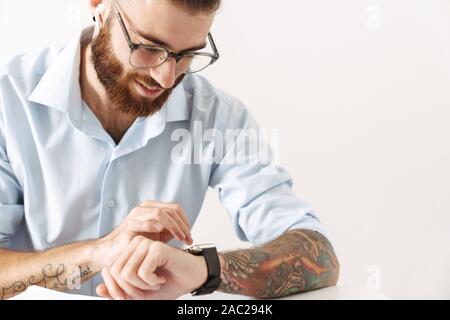 Image of happy young man wearing eyeglasses and earbuds looking at wristwatch while working at table in office Stock Photo