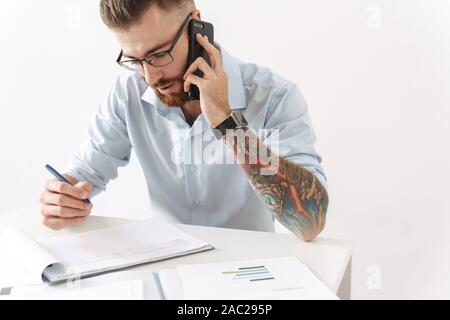 Image of concentrated young man wearing eyeglasses talking on smartphone while working in office Stock Photo