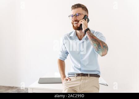 Image of delighted young man wearing eyeglasses talking on smartphone while working in office Stock Photo
