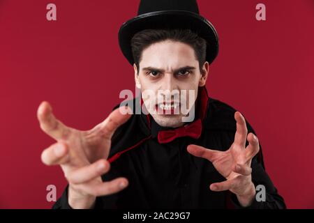 Image of vampire man with blood and fangs in black halloween costume attacking isolated over red wall Stock Photo