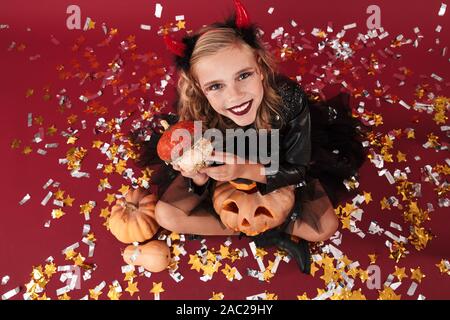 Picture of positive emotional optimistic little girl devil in carnival halloween costume isolated over red wall background holding pumpkin. Stock Photo