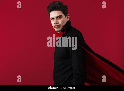 Image of serious vampire man with blood and fangs in black halloween costume looking at camera isolated over red wall Stock Photo