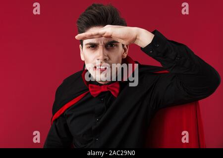 Image of handsome vampire man with blood and fangs in black halloween costume looking at camera isolated over red wall Stock Photo