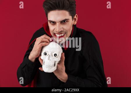 Image of vampire man with fangs in black halloween costume holding human skull isolated over red wall Stock Photo