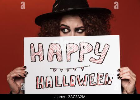 Image of scary witch girl in hat holding halloween text board isolated over red wall Stock Photo