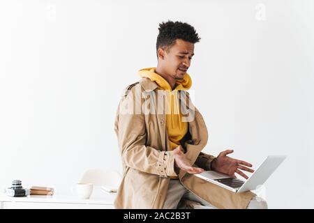 Image of perplexed african american guy wearing coat sitting with laptop in white studio Stock Photo