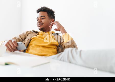 Image of smiling african american guy using smartphone and earbuds at table in white office Stock Photo