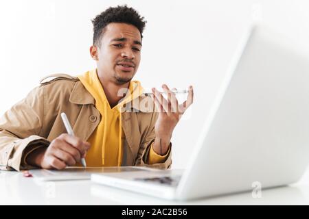 Image of perplexed african american guy using cellphone and tablet computer while working with laptop in white office Stock Photo